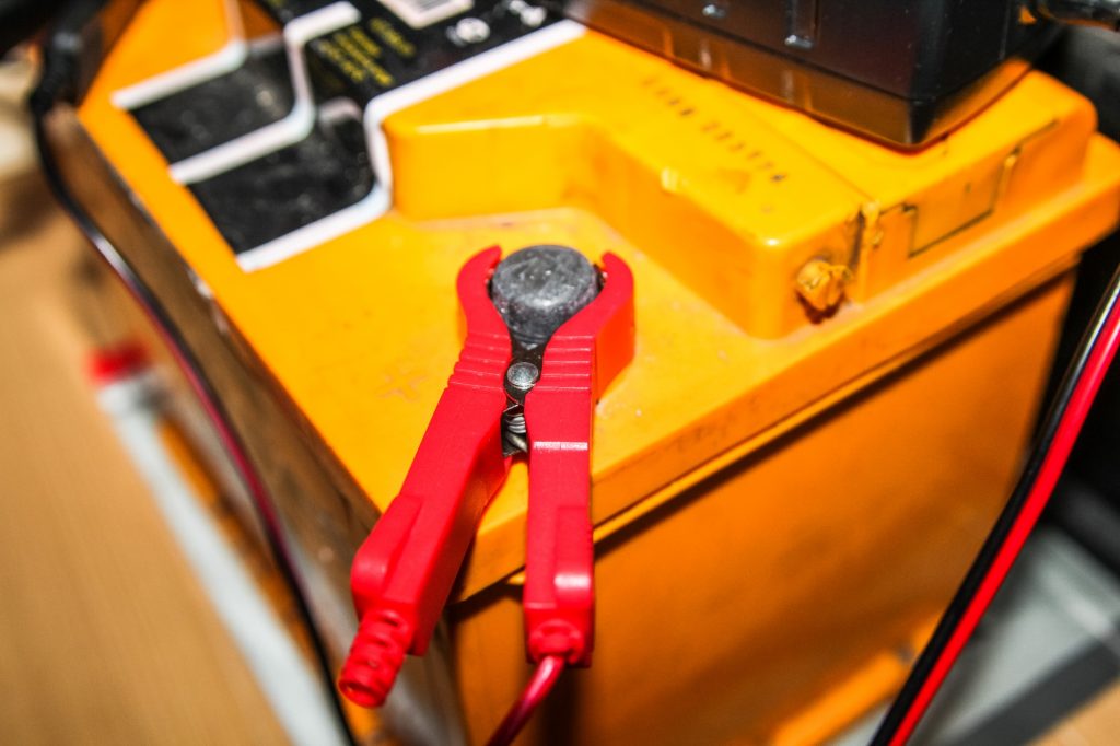The red positive jumper cable is attached to the positive terminal at the top of a yellow car battery.