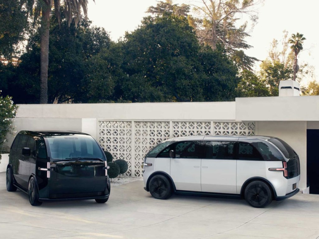 A white and a black Canoo electric vans sit in a driveway.