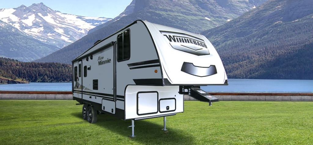 A white fifth wheel trailer is set in a snow-capped valley with a lake behind it.