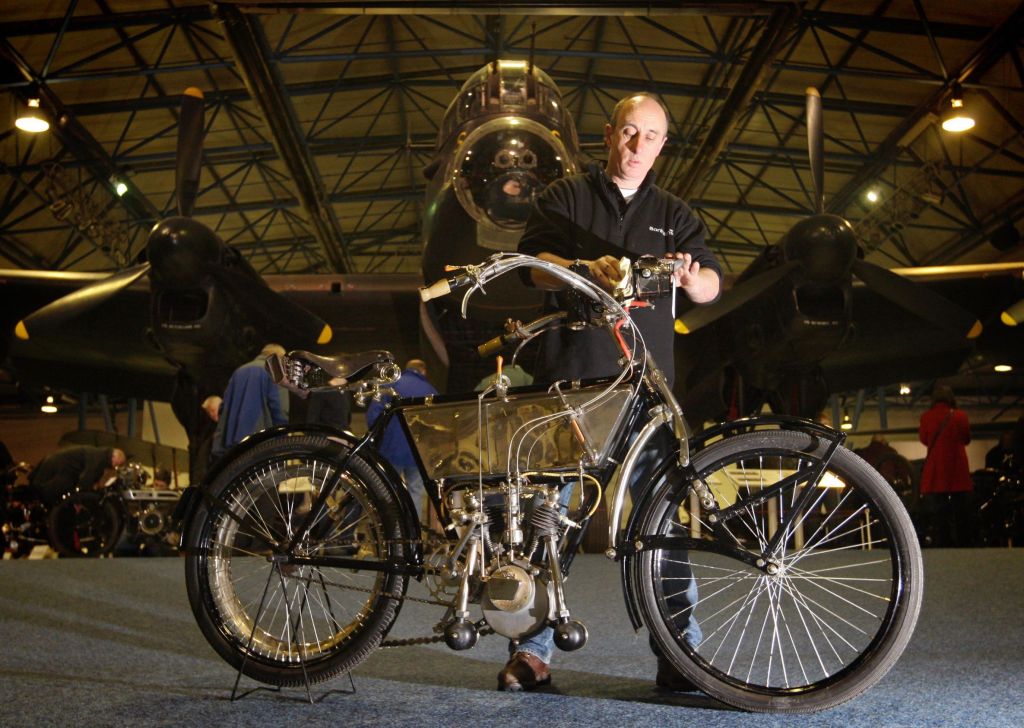 A 1905 Peugeot 3.5-hp V-twin motorcycle being photographed in front of an old airplane