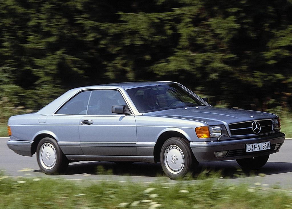 A silver-blue 1981 Mercedes S-Class Coupe drives down a forested road
