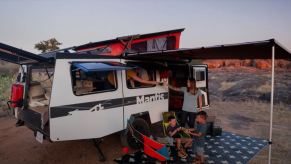 A white trailer RV with the pop-up extended sits on a trial with a family sitting under the exterior awning.