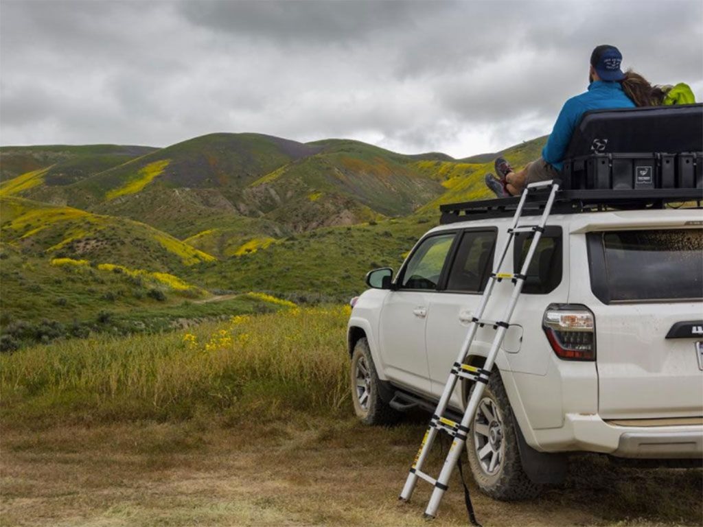 A man sits on the roof rack of a white Toyota 4Runner with a Front Runner aluminum telescopic ladder