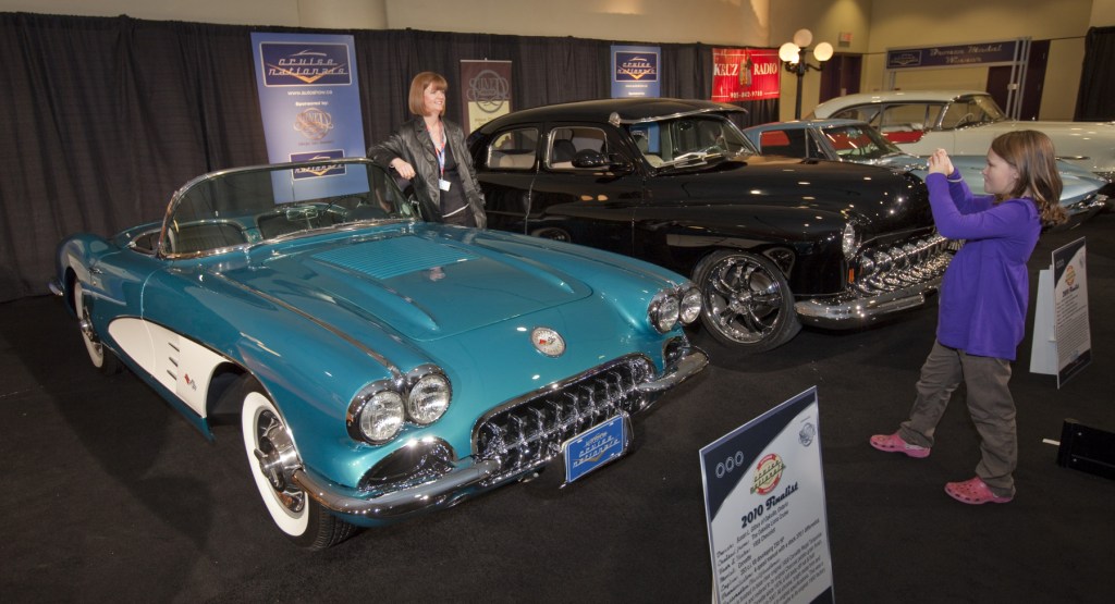 A light blue 1958 Chevrolet Corvette C1 displayed at an auto show. Two other classic cars are pictured to the right of it.