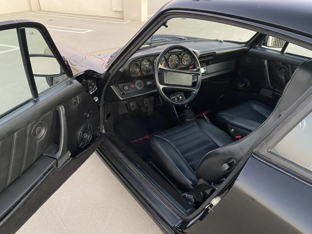 The front interior and dashboard of a black 1982 Porsche 911 SC seen through the open driver's door