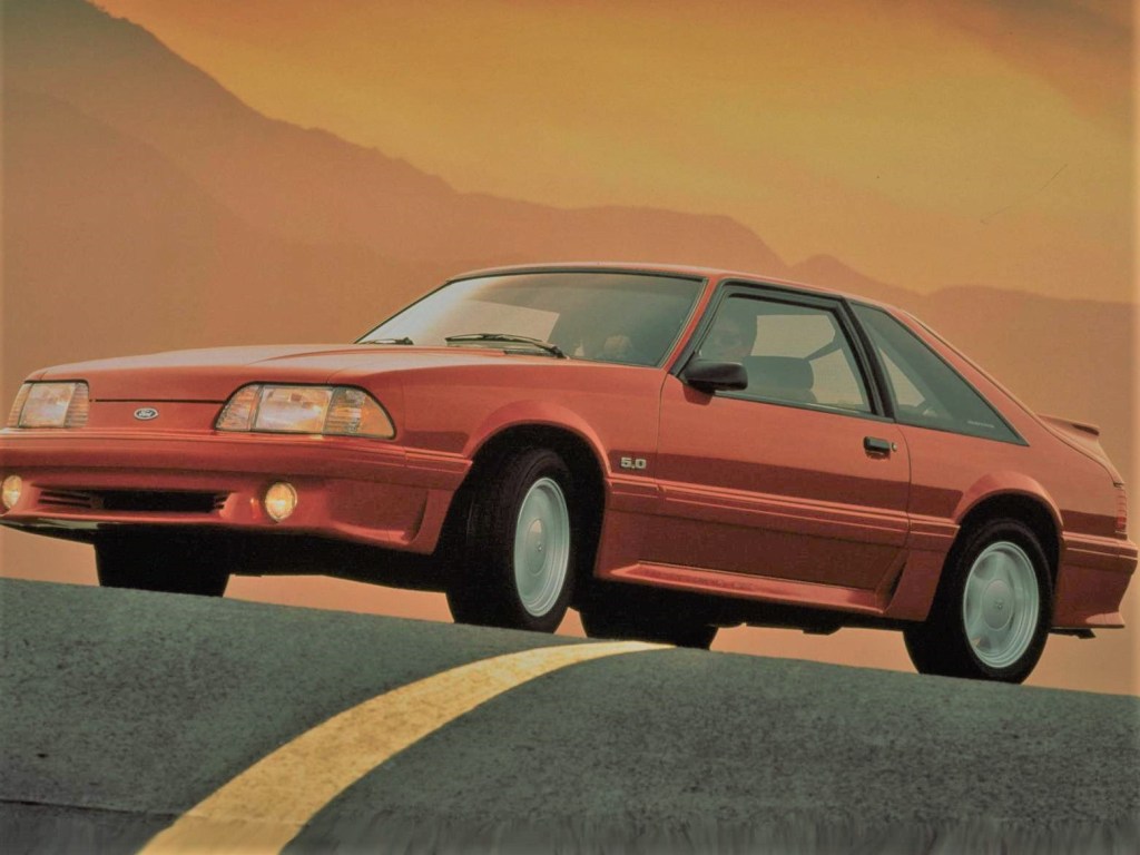 A red Ford Mustang GT in the foreground against a sunrise across a mountain road.