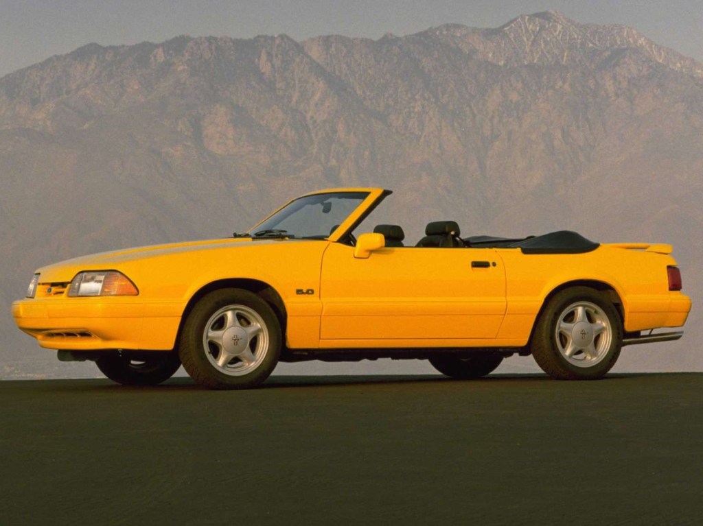 A yellow, convertible 1993 For Mustang sits at the base of a mountain with its top down. 