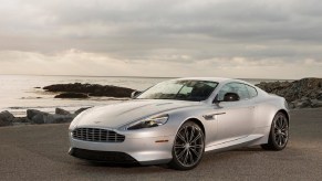 A silver Aston Martin DB9 sits on the side of a coastal road.