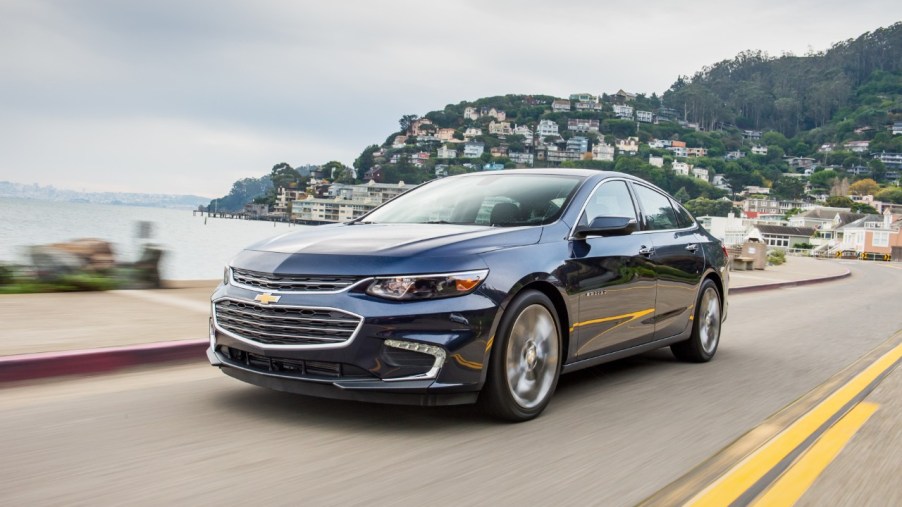 The 2018 Chevy Malibu driving down the road with the ocean and a town in the background.