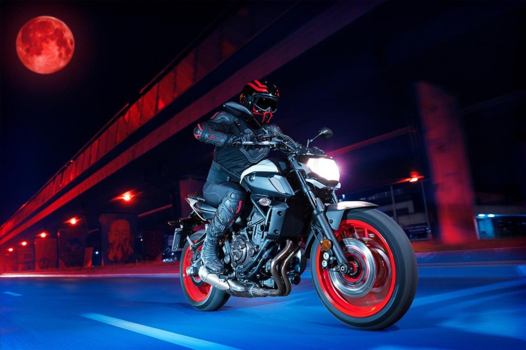 A black-clad rider rides a white 2020 Yamaha MT-07 through a stylized night cityscape