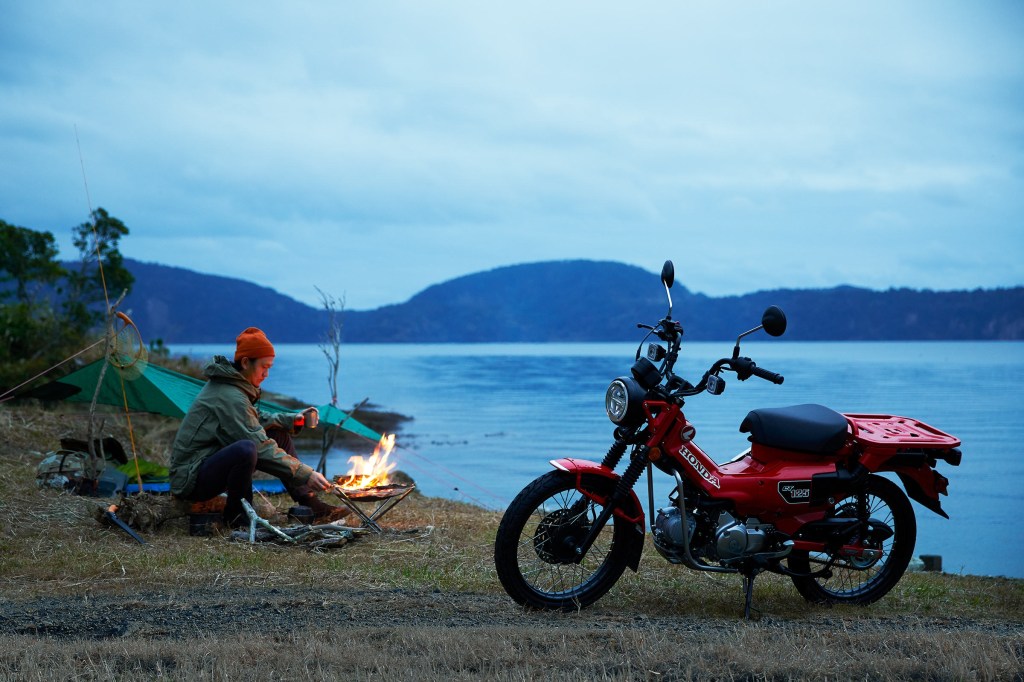 A red 2021 Honda Trail125 ABS by a lake and a campsite