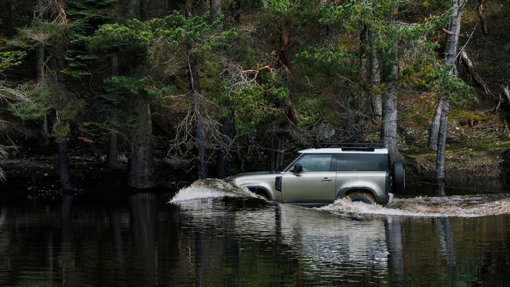 A green 2021 Land Rover Defender 90 wades through a forest river