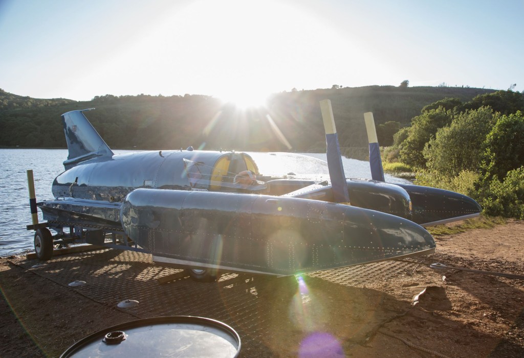 Donald Campbell's blue Bluebird hydroplane rests on its stand in front of a sunlit Scottish lake