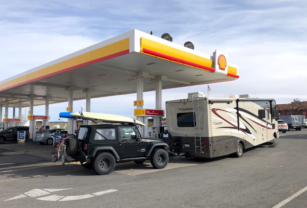 An RV tows a Jeep and outdoors gear while filling up its gas tank