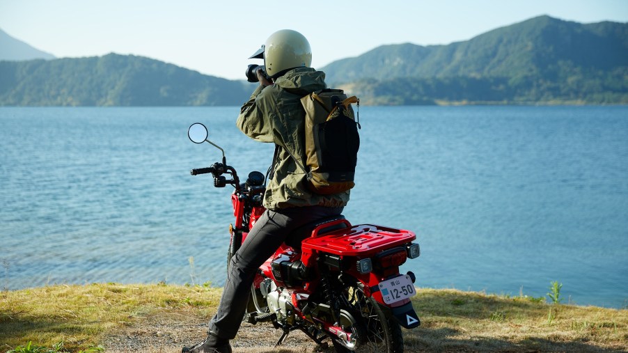 A rider sits on a red 2021 Honda Trail 125 by a lake
