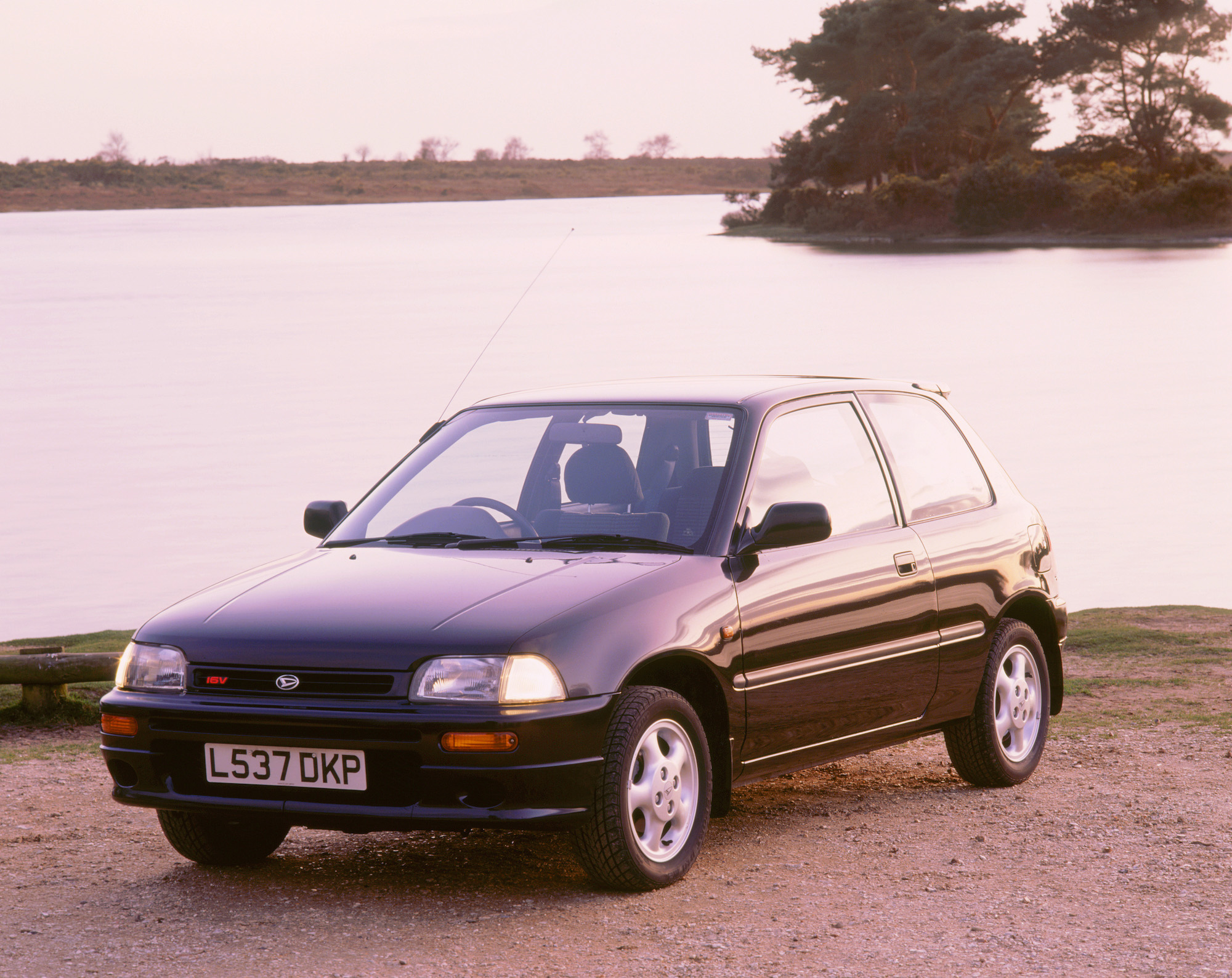 A dark-colored 1994 Daihatsu Charade GTI hatchback parked by a lake