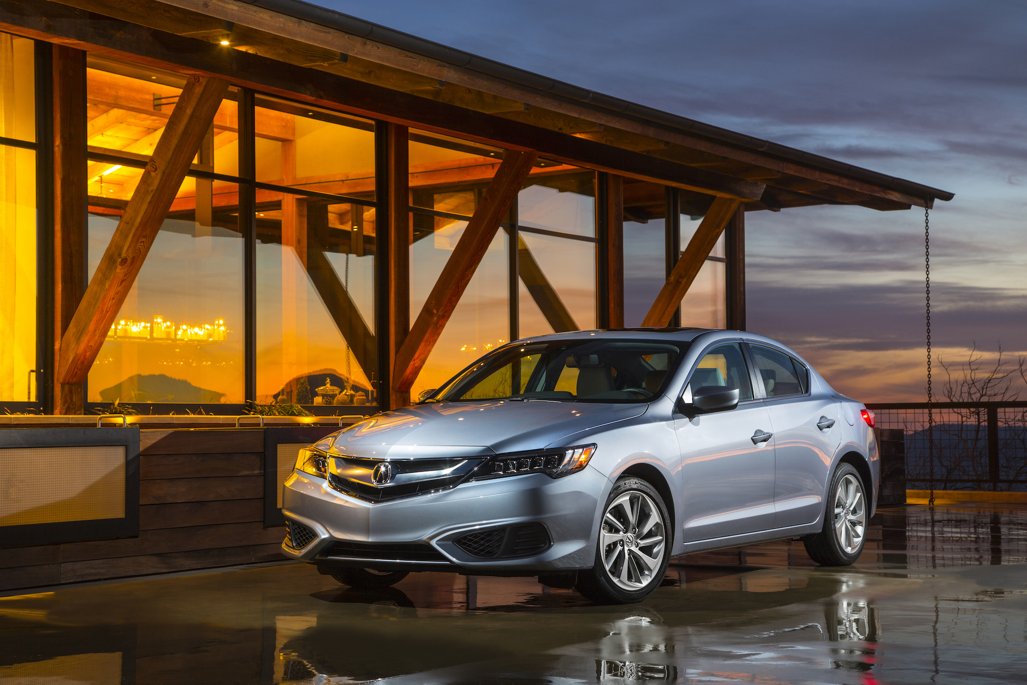 A silver 2016 Acura ILX parked in front of a wood building with large windows and a sunset in the background