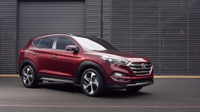 A dark-red 2016 Hyundai Tucson parked on pavement in front of a dark-gray building