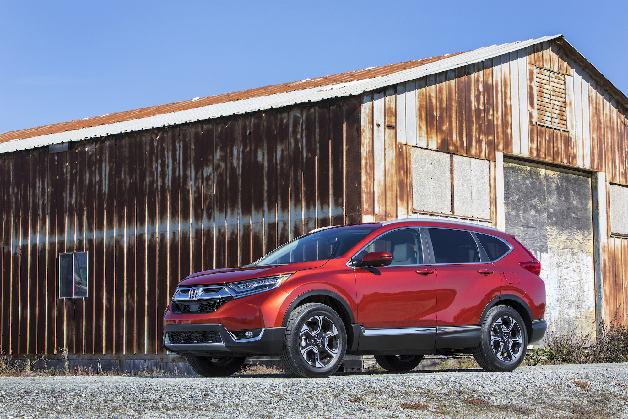 A metallic red 2018 Honda CR-V parked on gravel in front of a rusty barn and blue sky