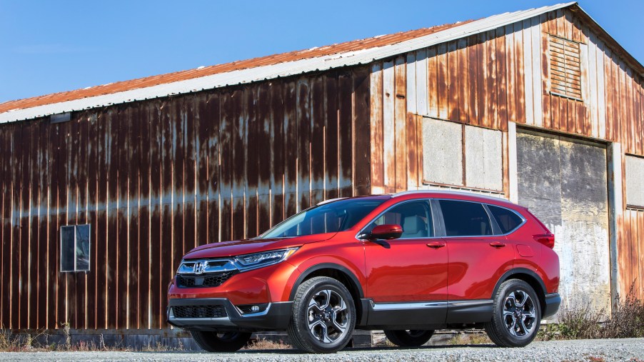 A metallic red 2018 Honda CR-V parked on gravel in front of a rusty barn and blue sky