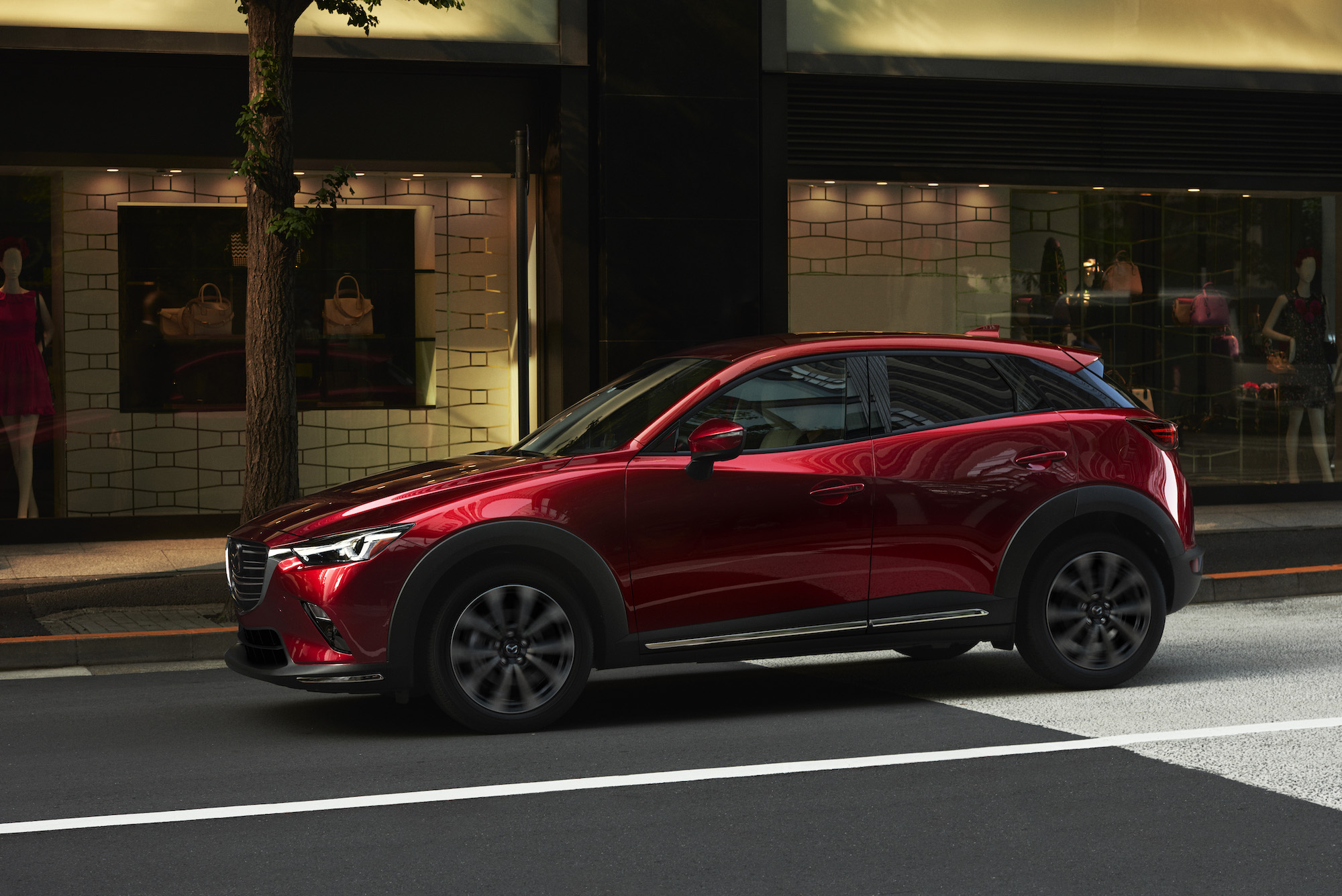 A metallic red 2019 Mazda CX-3 drives on a city street outside a storefront
