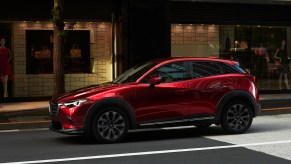 A metallic red 2019 Mazda CX-3 drives on a city street outside a storefront
