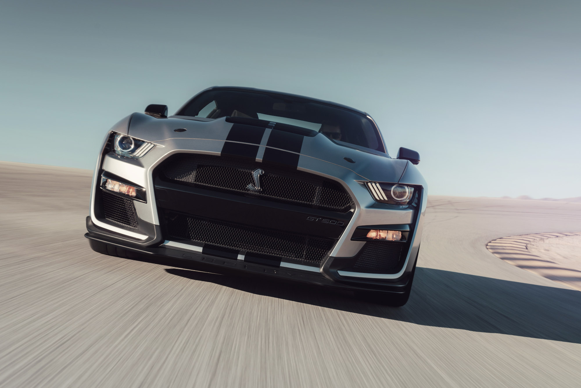 A 2020 Ford Mustang Shelby GT500 with a silver paint job and black racing stripes drives on a racetrack in the desert