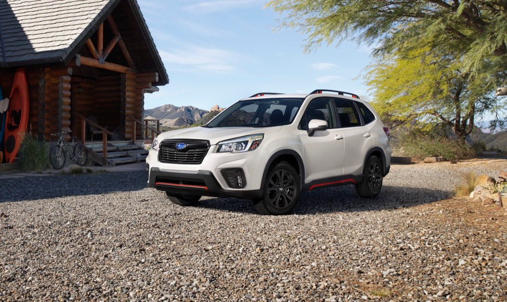A White Subaru Forester is parked on gravel in front of a log cabin in the mountains.