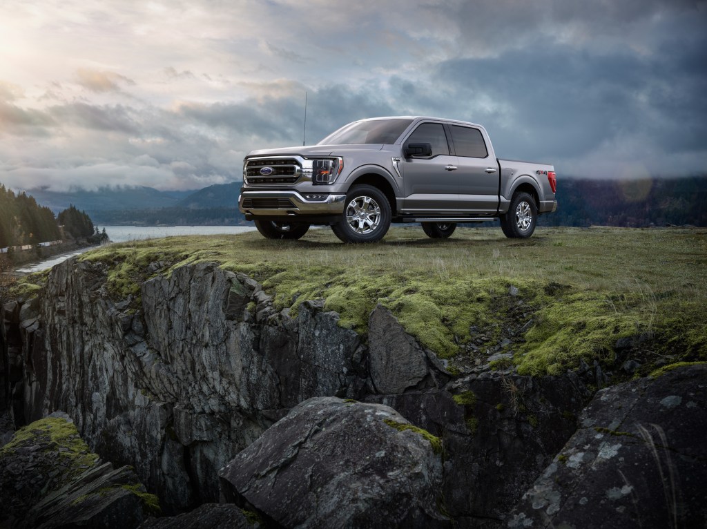 A 2021 Ford F-150 Platinum pickup truck in Iconic Silver stands atop a grassy cliff overlooking water and pine trees