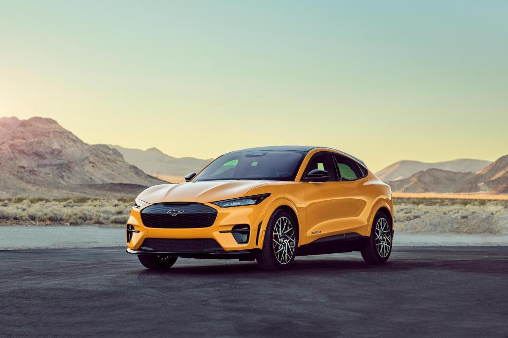 A bright-yellow 2021 Ford Mustang Mach-E GT Performance Edition compact SUV is parked in front of a lake surrounded by mountains on a clear day.