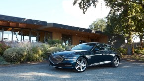 A dark-colored 2021 Genesis G80 parked in the shade of a tree outside a midcentury modern home