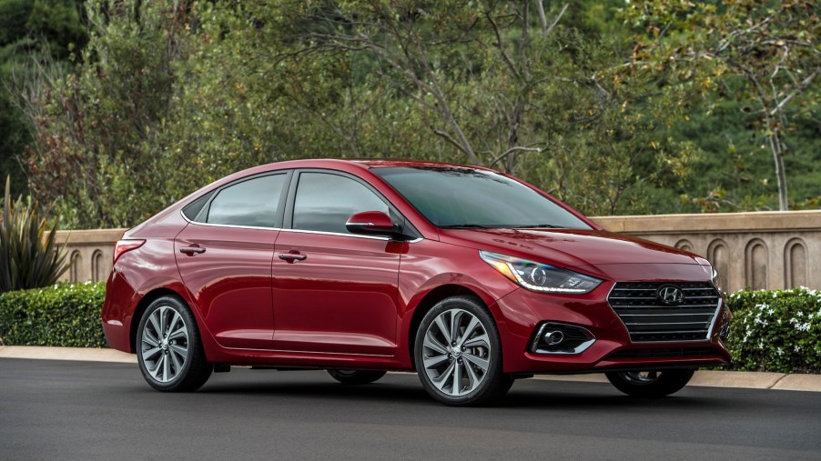 A dark-red 2021 Hyundai Accent is parked on pavement in front of foliage and a concrete wall