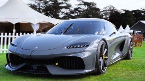 A gray Koenigsegg Gemera supercar is parked on a green lawn with a white tent in the background.