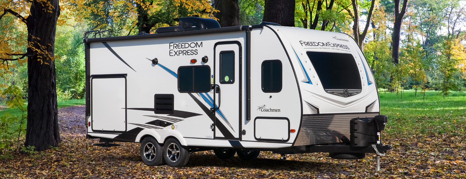 A double axle travel trailer sits in the leaves by a tree.