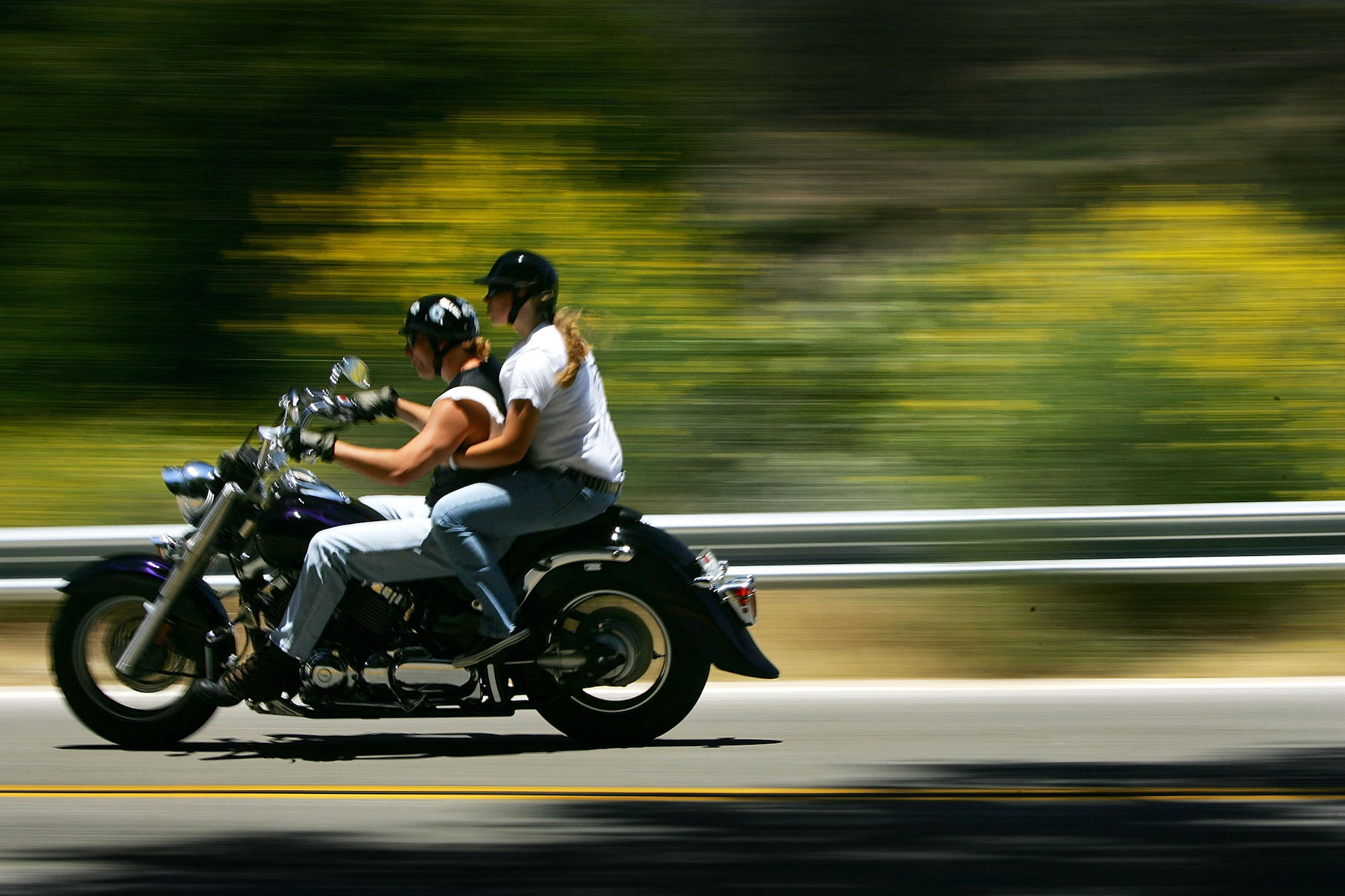 A motorcycle with two riders drives past wildflowers on the side of a road