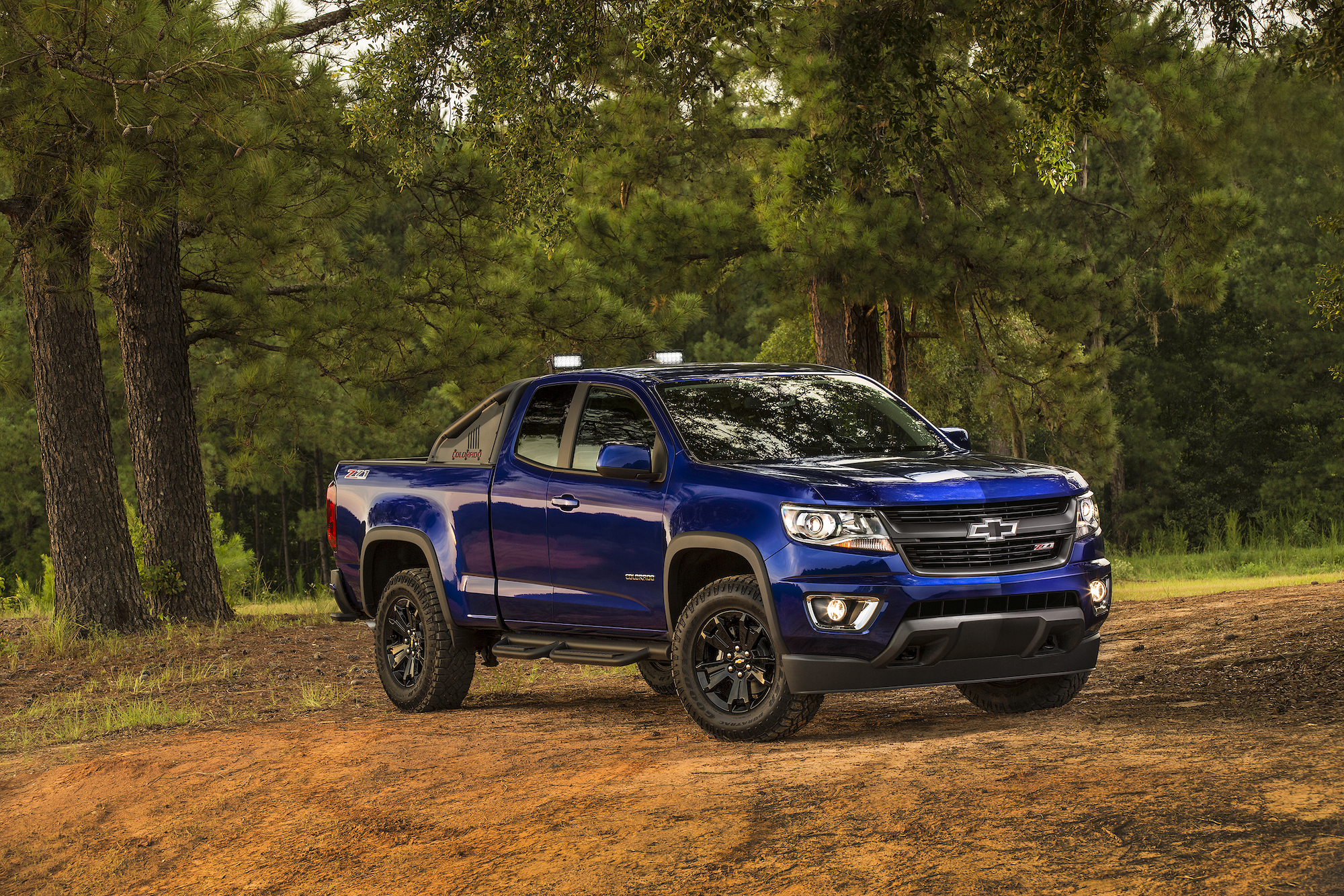 A dark-blue 2016 Chevy Colorado Trail Boss parked on red soil in front of shrubbery and trees