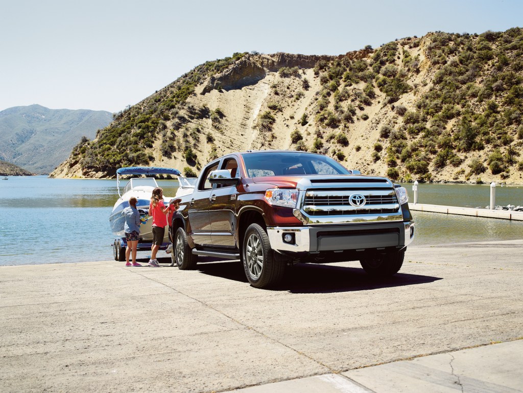 Two children stand next to a maroon 2016 Toyota Tundra towing a boat on a boat ramp overlooking a lake and mountains
