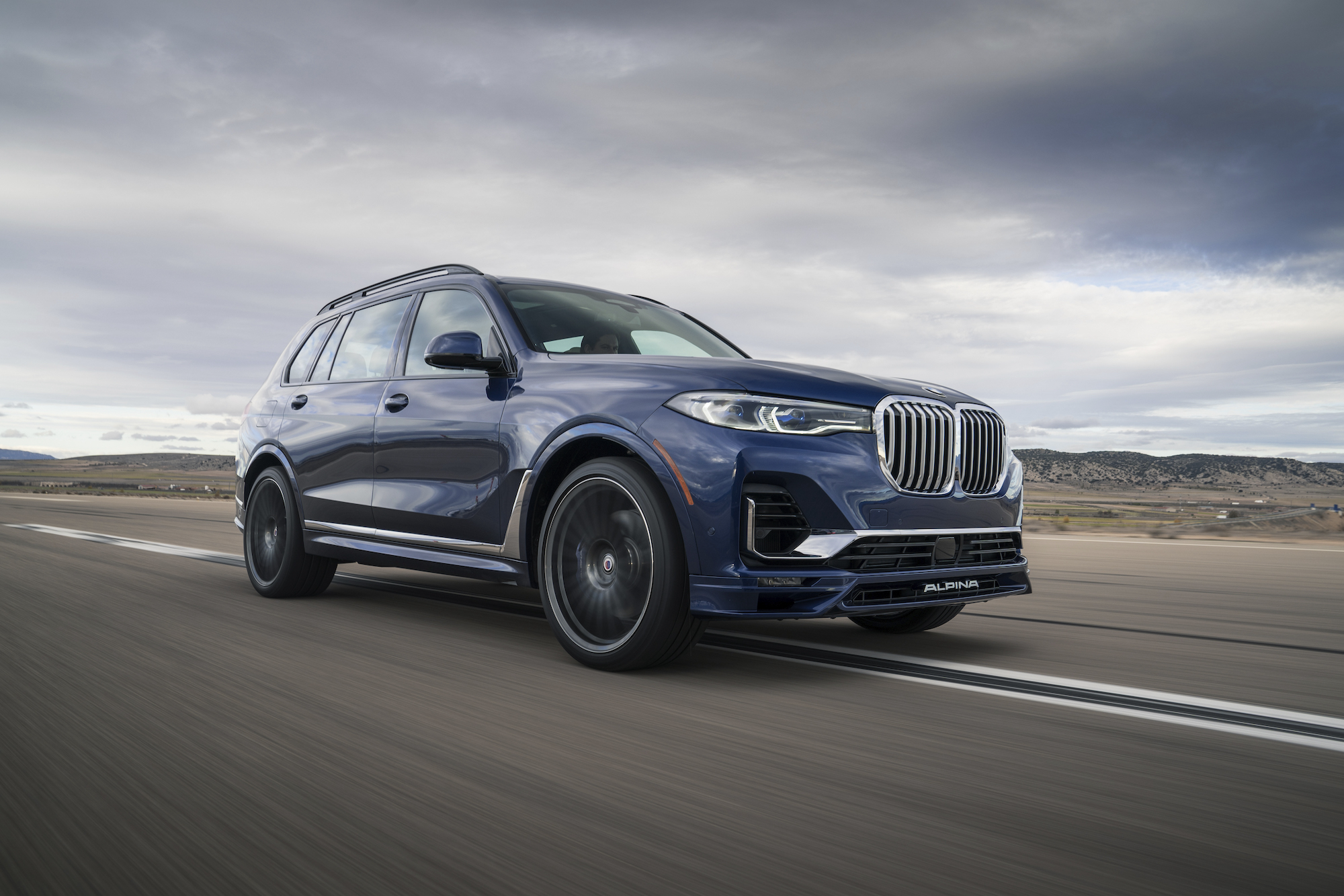 A dark-blue 2021 BMW Alpina XB7 large crossover SUV travels on a strip of asphalt surrounded by grassy hills