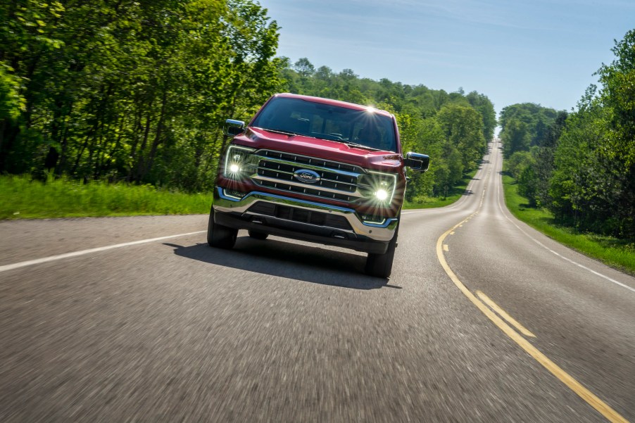 A red 2021 Ford F-150 Lariat in Rapid Red Metallic Tinted Clearcoat travels on a two-lane highway flanked by foliage