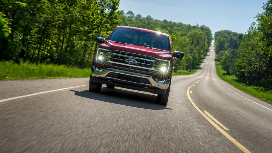 A red 2021 Ford F-150 Lariat in Rapid Red Metallic Tinted Clearcoat travels on a two-lane highway flanked by foliage