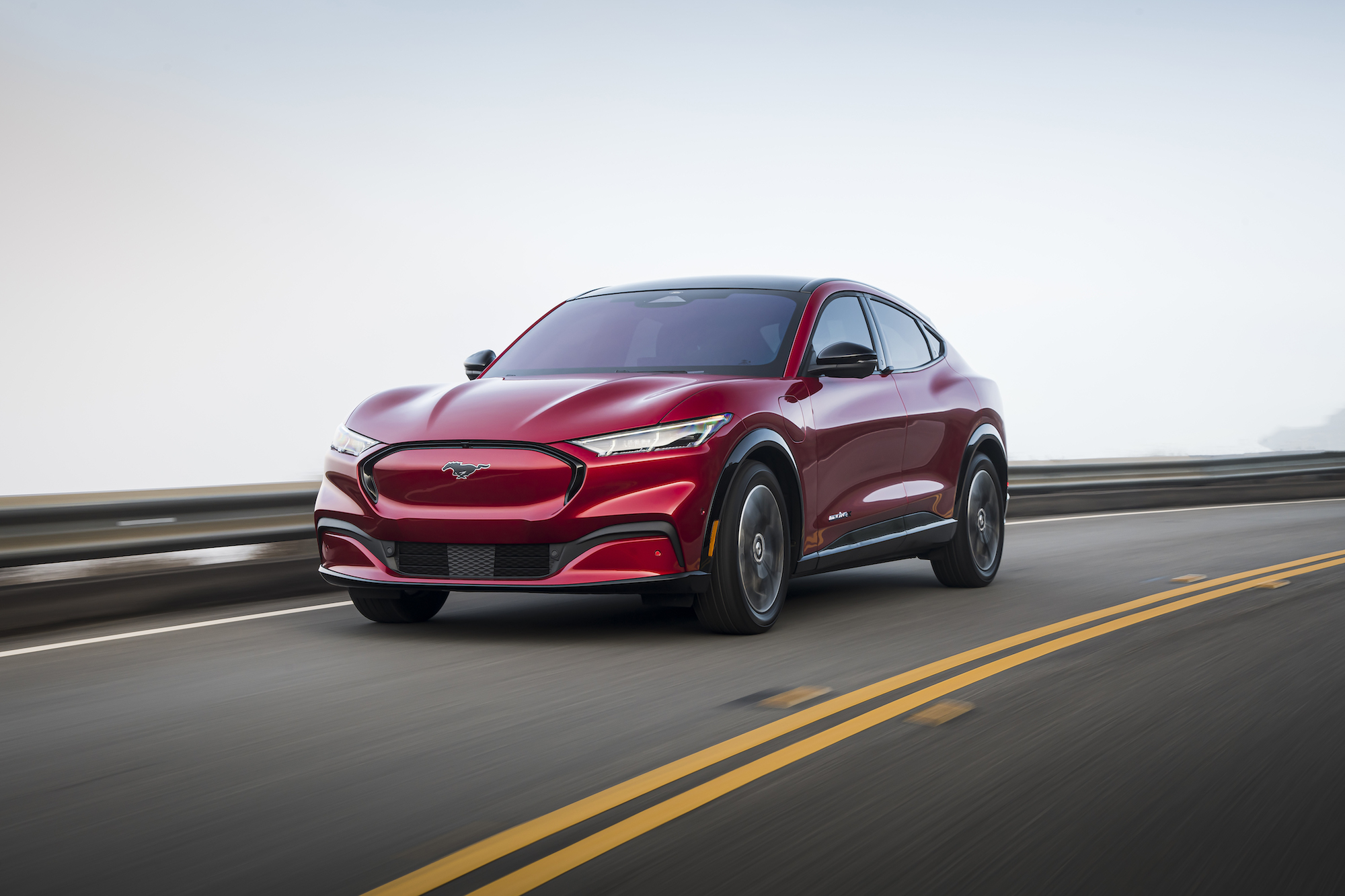A red 2021 Ford Mustang Mach-E travels on a paved two-lane road along a guardrail