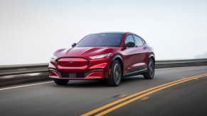 A red 2021 Ford Mustang Mach-E travels on a paved two-lane road along a guardrail