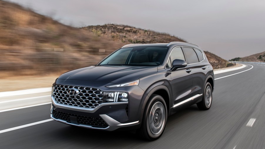 A dark-gray 2021 Hyundai Santa Fe travels on a two-lane highway flanked by hills on a cloudy day