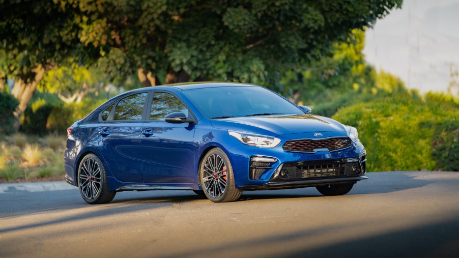 A blue 2021 Kia Forte GT Sport compact sedan parked on a paved road in front of green foliage