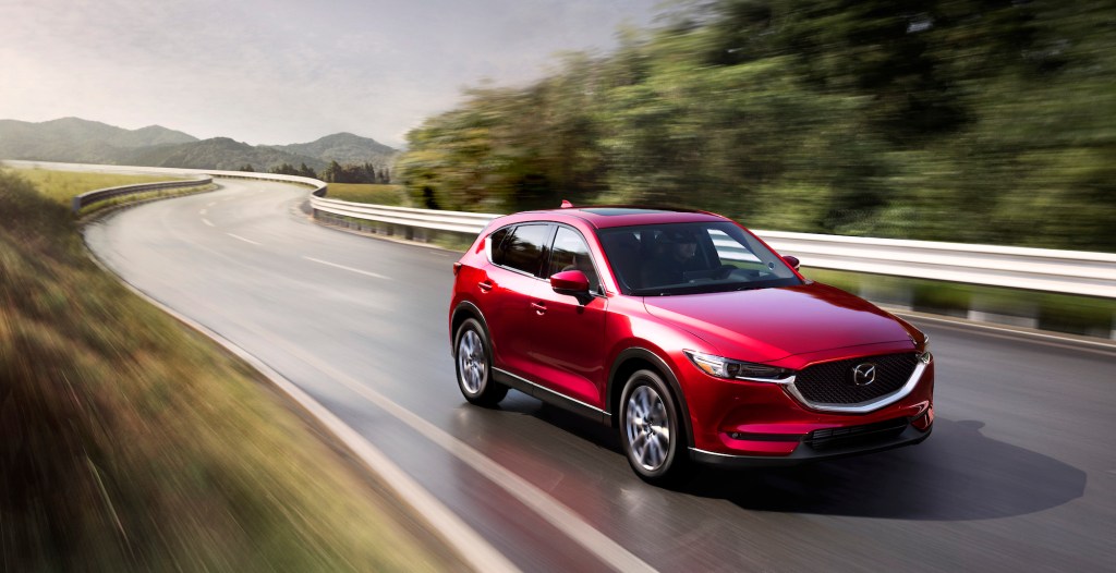 A metallic red 2021 Mazda CX-5 travels on a wet two-lane highway lined by metal guardrails and green mountains