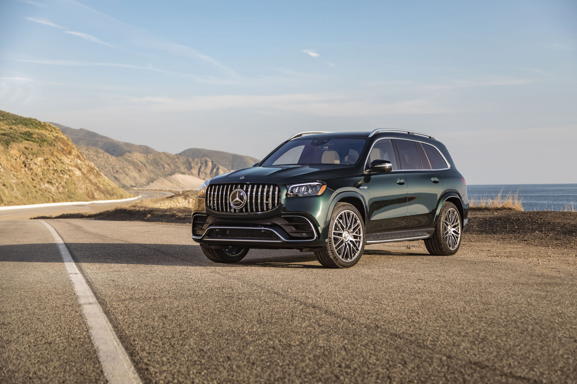 A dark-green 2021 Mercedes-AMG GLS 63 parked on a paved road with blue sky and mountains in the background