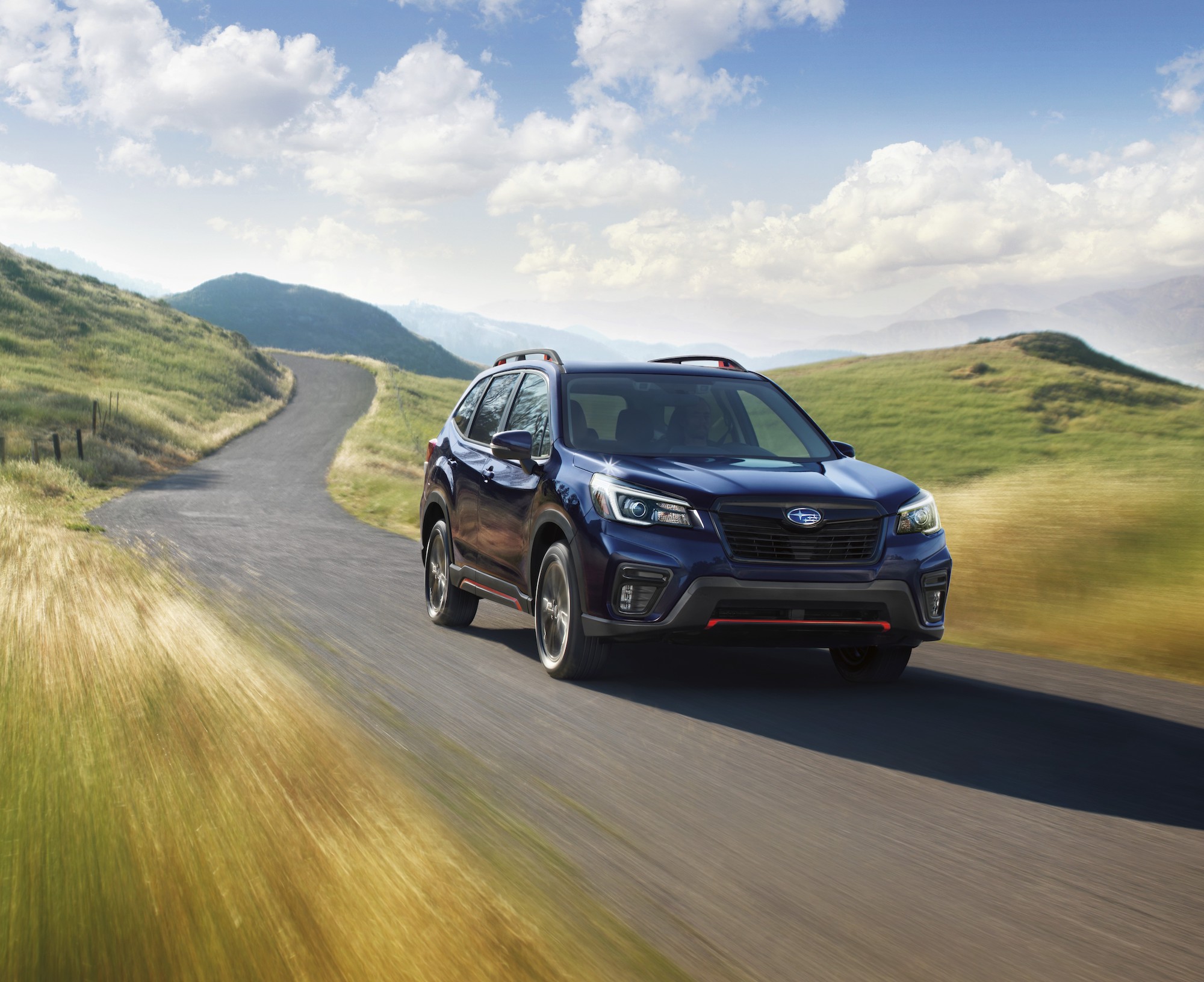 A dark-colored 2021 Subaru Forester travels on a paved road through grassy hills