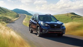 A dark-colored 2021 Subaru Forester travels on a paved road through grassy hills