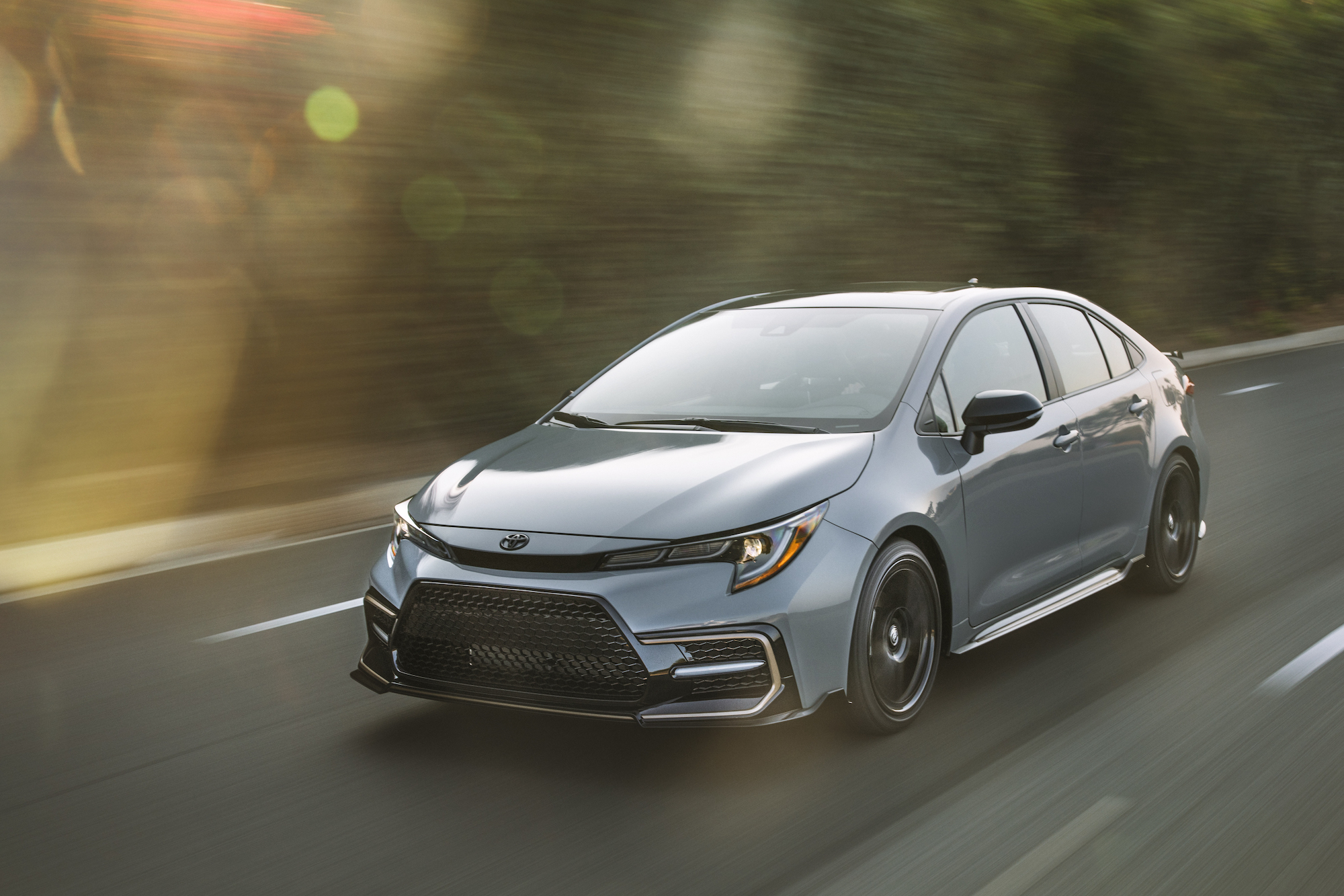 A silver 2021 Toyota Corolla Apex Edition travels on a two-lane paved road lined by foliage
