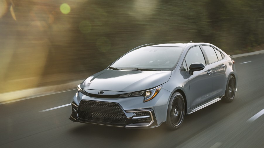 A silver 2021 Toyota Corolla Apex Edition travels on a two-lane paved road lined by foliage
